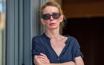 Woman with blonde hair in a bun, wearing dark sunglasses and a navy blue shirt, stands against a door frame with arms crossed, looking serious.