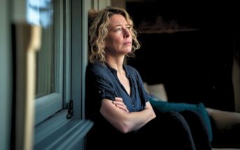 A woman with curly hair sits indoors by a window, arms crossed, gazing outside with a thoughtful and somber expression. The room is dimly lit, emphasizing her reflective mood.