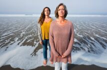 Two women stand barefoot on a sandy beach near the shoreline, with waves approaching. The woman in front wears a light pink sweater and white pants, while the woman behind her wears a yellow top and gray cardigan.