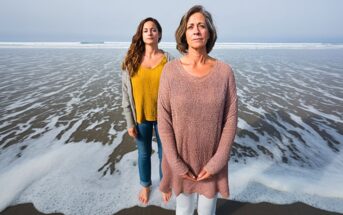 Two women stand barefoot on a sandy beach near the shoreline, with waves approaching. The woman in front wears a light pink sweater and white pants, while the woman behind her wears a yellow top and gray cardigan.
