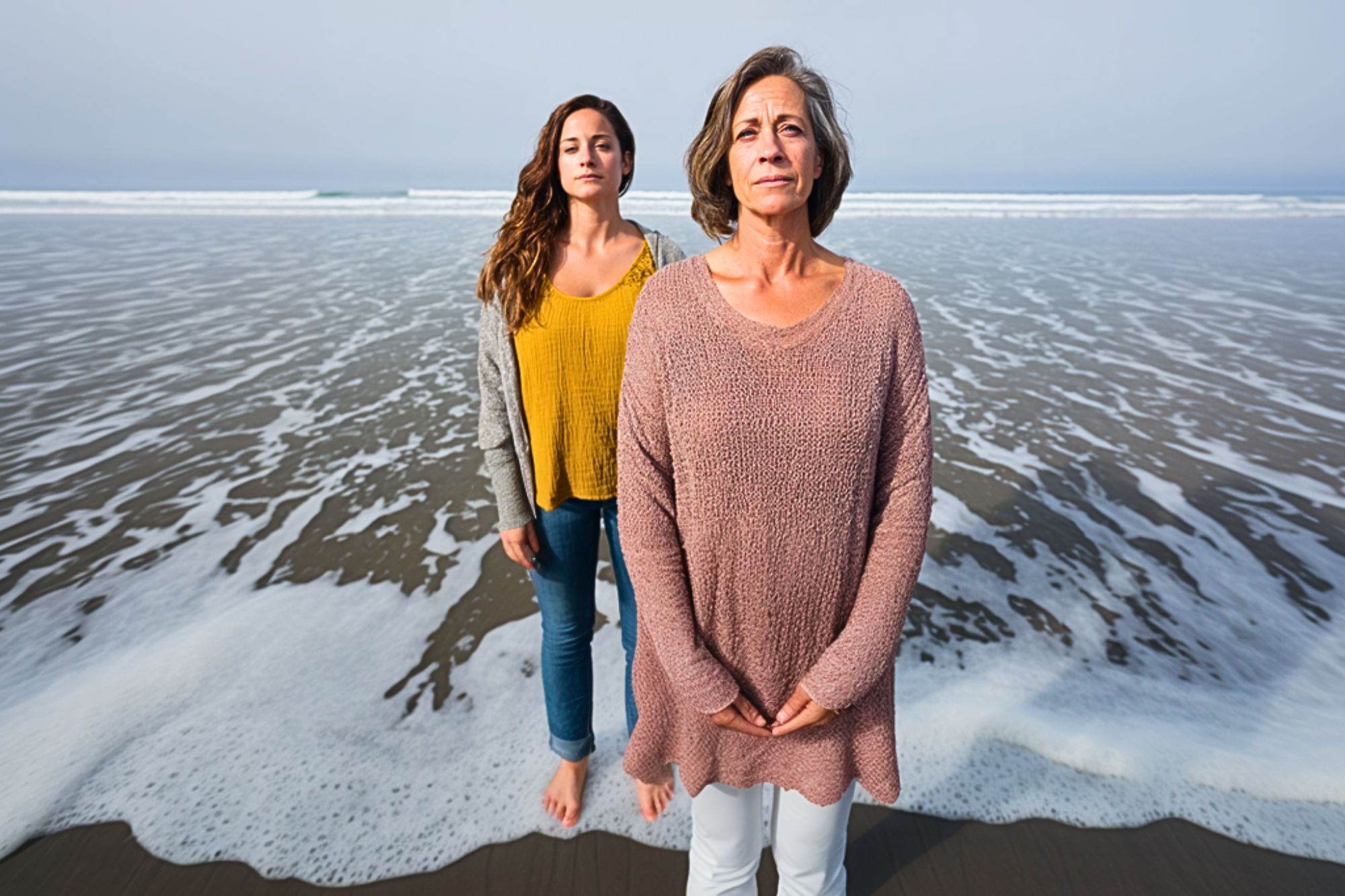 Two women stand barefoot on a sandy beach near the shoreline, with waves approaching. The woman in front wears a light pink sweater and white pants, while the woman behind her wears a yellow top and gray cardigan.