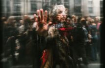 An older woman with gray hair and a red scarf touches a glass window, looking contemplative. Blurred reflections and people in the background create a sense of movement and separation.