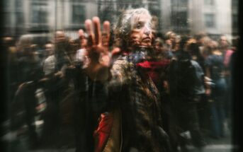 An older woman with gray hair and a red scarf touches a glass window, looking contemplative. Blurred reflections and people in the background create a sense of movement and separation.