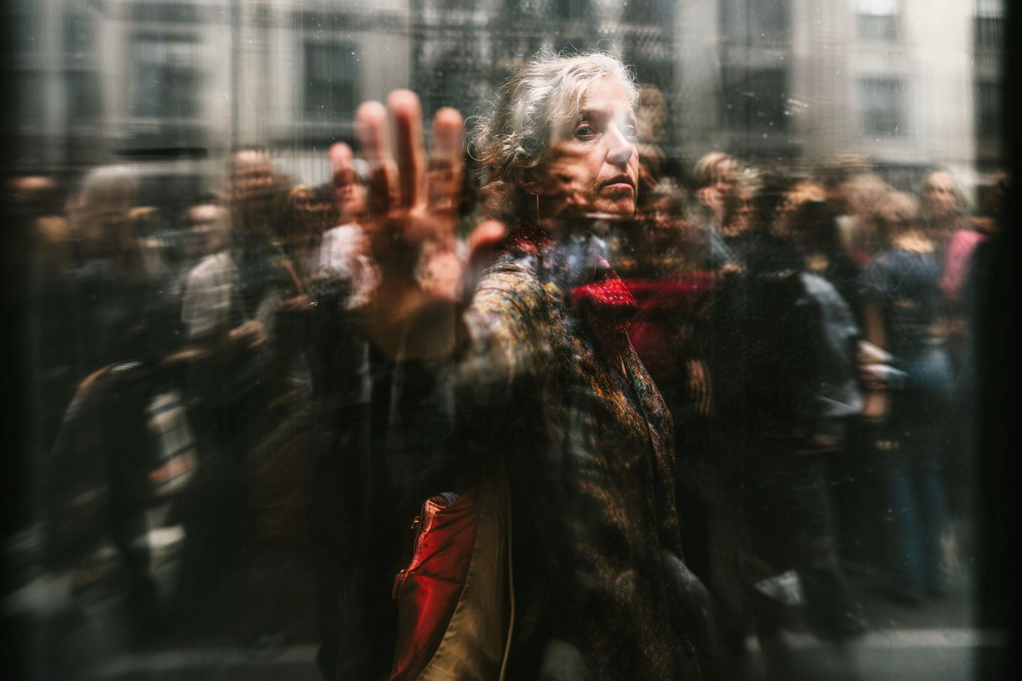 An older woman with gray hair and a red scarf touches a glass window, looking contemplative. Blurred reflections and people in the background create a sense of movement and separation.