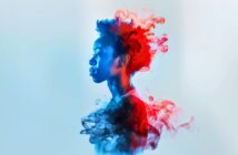 A side profile of a young person with an afro, illuminated by blue and red light. Colorful, smoky clouds in blue and red swirl around and behind their head and shoulders against a pale background.