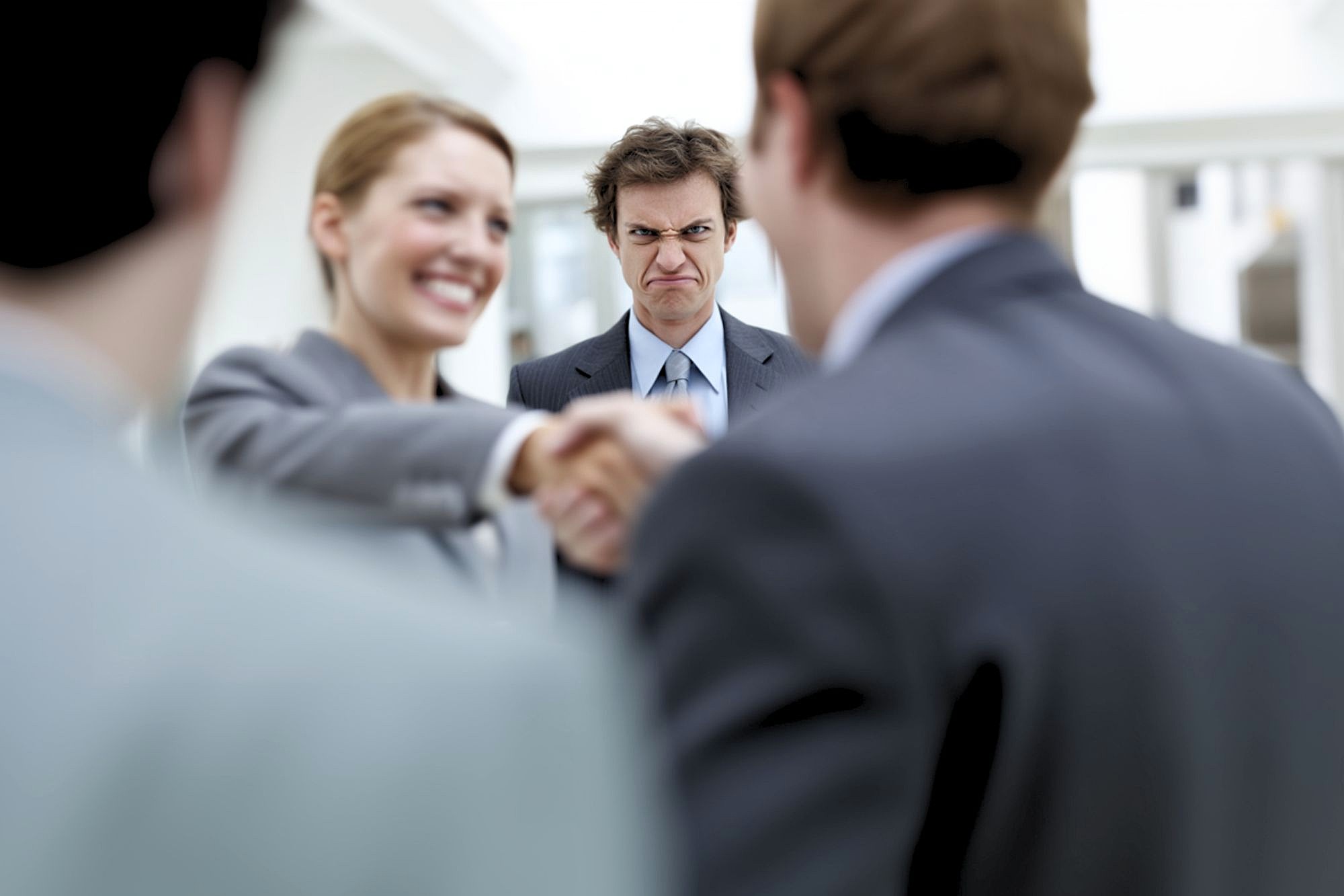 A man in the background looks upset while two people in the foreground smile and shake hands, with another person partially visible to the side.