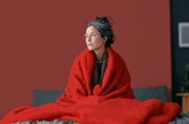 A woman with glasses and gray hair sits on a bed wrapped in a large red blanket, looking to the side. The background is a plain red wall with part of a picture frame visible.