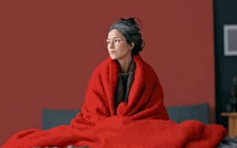 A woman with glasses and gray hair sits on a bed wrapped in a large red blanket, looking to the side. The background is a plain red wall with part of a picture frame visible.