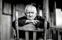 An elderly man with white hair sits on a wooden chair, resting his folded arms on its back, gazing directly at the camera with a thoughtful expression. The photo is black and white, adding a somber tone.