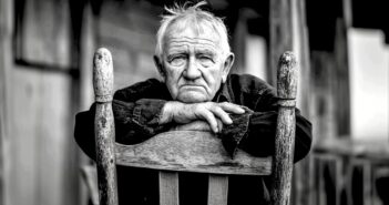 An elderly man with white hair sits on a wooden chair, resting his folded arms on its back, gazing directly at the camera with a thoughtful expression. The photo is black and white, adding a somber tone.