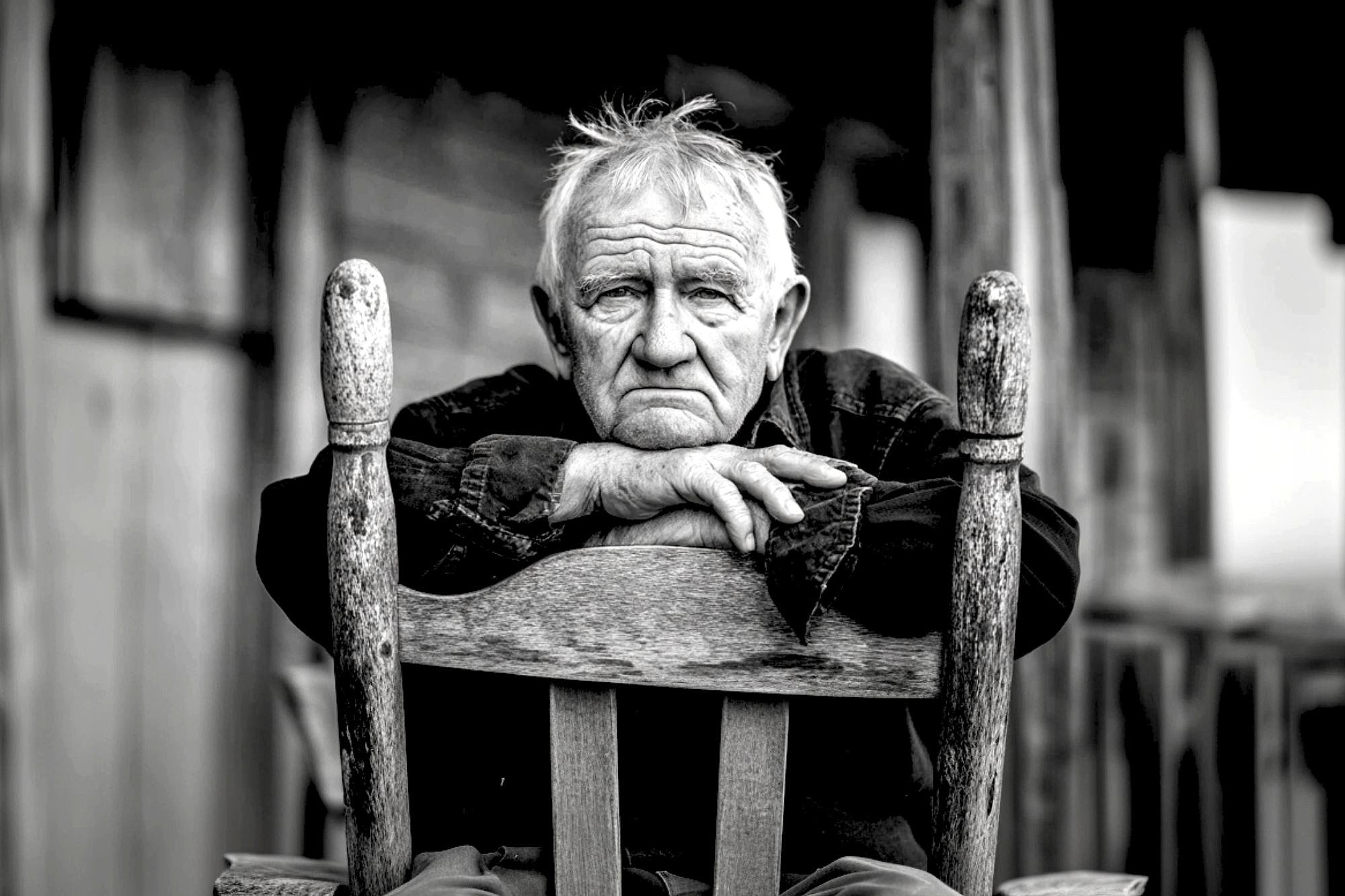 An elderly man with white hair sits on a wooden chair, resting his folded arms on its back, gazing directly at the camera with a thoughtful expression. The photo is black and white, adding a somber tone.