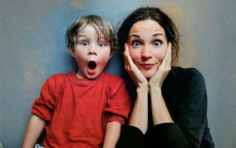 A child in a red shirt and an adult in black sit side by side, both making surprised facial expressions with wide eyes. The adult is resting their face on their hands.