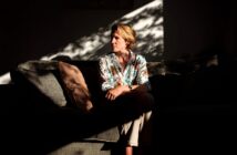 A woman in a floral shirt sits on a sofa in a dim room, illuminated by sunlight streaming through a window, creating shadows and patterns on the wall and her face.