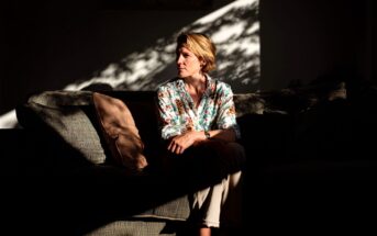 A woman in a floral shirt sits on a sofa in a dim room, illuminated by sunlight streaming through a window, creating shadows and patterns on the wall and her face.