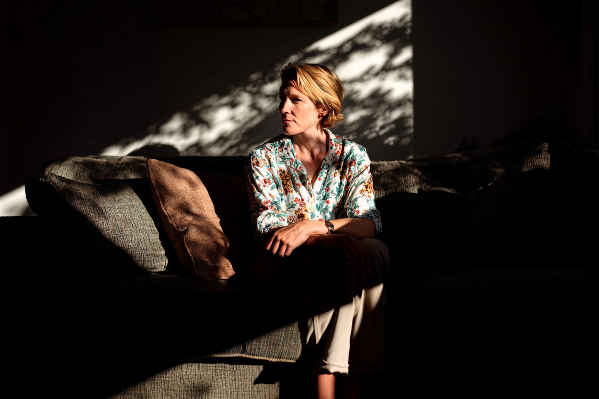 A woman in a floral shirt sits on a sofa in a dim room, illuminated by sunlight streaming through a window, creating shadows and patterns on the wall and her face.