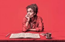 A woman sits at a table with a map, looking thoughtfully upward. She rests her chin on her hand and wears a red shirt. On the table are a pen, a notebook, and a cup of coffee. The background is solid red.