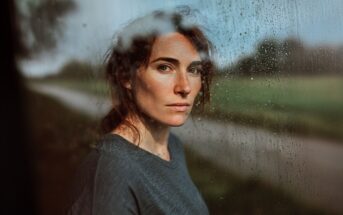 A woman with loose brown hair stands by a rain-speckled window, looking directly at the camera. The blurred green landscape is visible outside, and natural light highlights her face.