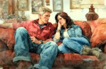 A man and woman sit close together on a red couch, casually dressed in denim and relaxed poses, looking at each other in a cozy, warmly lit living room with framed pictures and a lamp in the background.