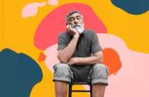 An older man with a white beard sits on a stool, resting his chin on his hand, looking thoughtful. He is in front of a vibrant yellow background with abstract pink, orange, and blue shapes.