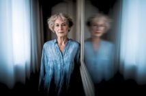 An older woman with short, curly gray hair stands indoors, wearing a blue shirt. She leans against a wall with her reflection blurred in a mirror beside her, giving a dreamy, contemplative atmosphere.