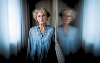 An older woman with short, curly gray hair stands indoors, wearing a blue shirt. She leans against a wall with her reflection blurred in a mirror beside her, giving a dreamy, contemplative atmosphere.