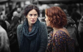 Two women are talking outdoors in a crowd. The woman on the left, with dark hair and a scarf, looks at the other woman, who has curly red hair and wears a flower-patterned top. The background is blurred in black and white.