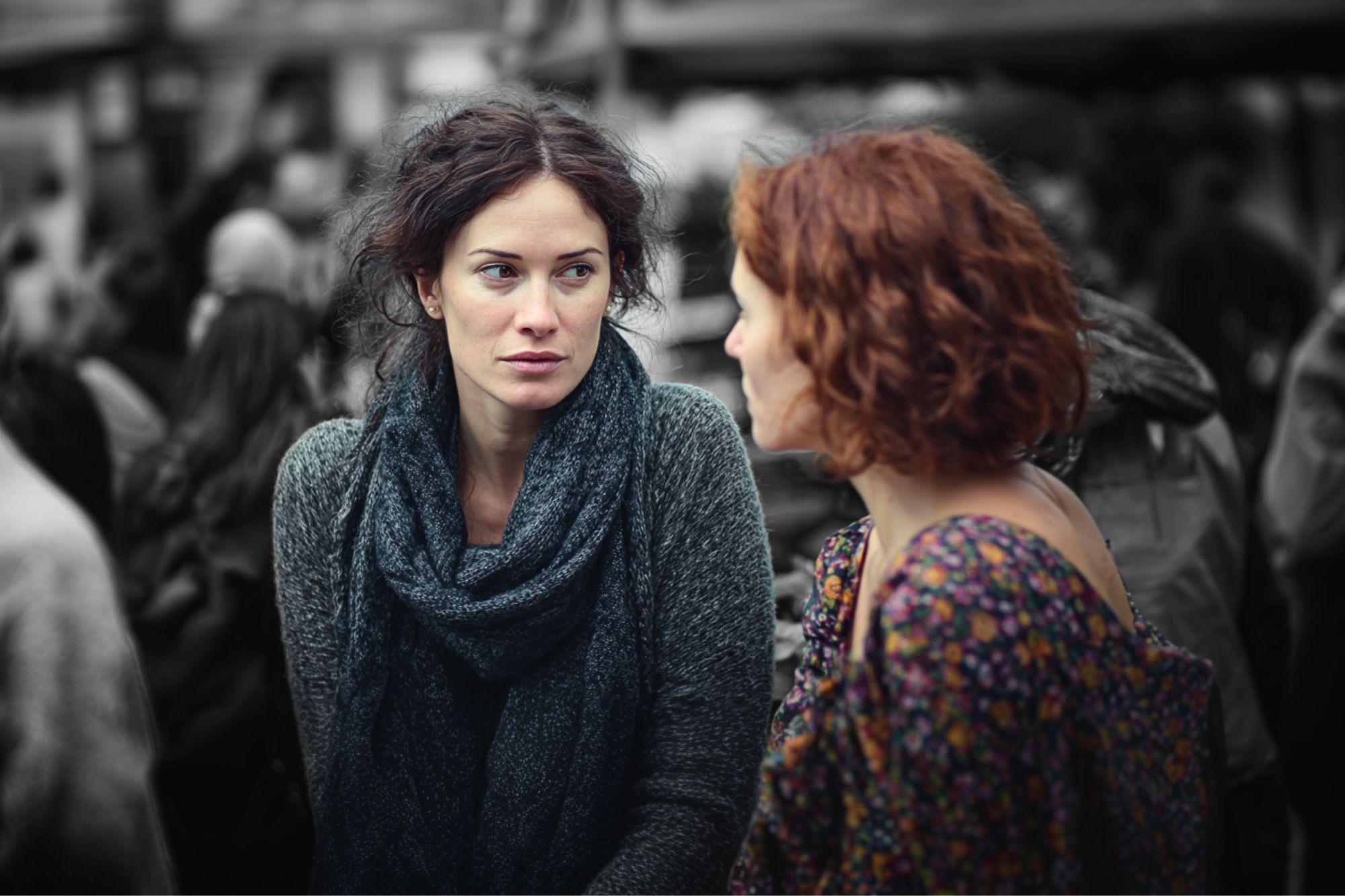 Two women are talking outdoors in a crowd. The woman on the left, with dark hair and a scarf, looks at the other woman, who has curly red hair and wears a flower-patterned top. The background is blurred in black and white.