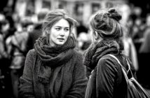 Two young women with scarves talk to each other outdoors in a crowded area. The image is black and white, with a blurred background of people, emphasizing their conversation and expressions.