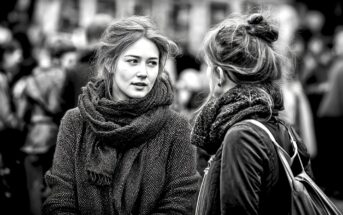 Two young women with scarves talk to each other outdoors in a crowded area. The image is black and white, with a blurred background of people, emphasizing their conversation and expressions.