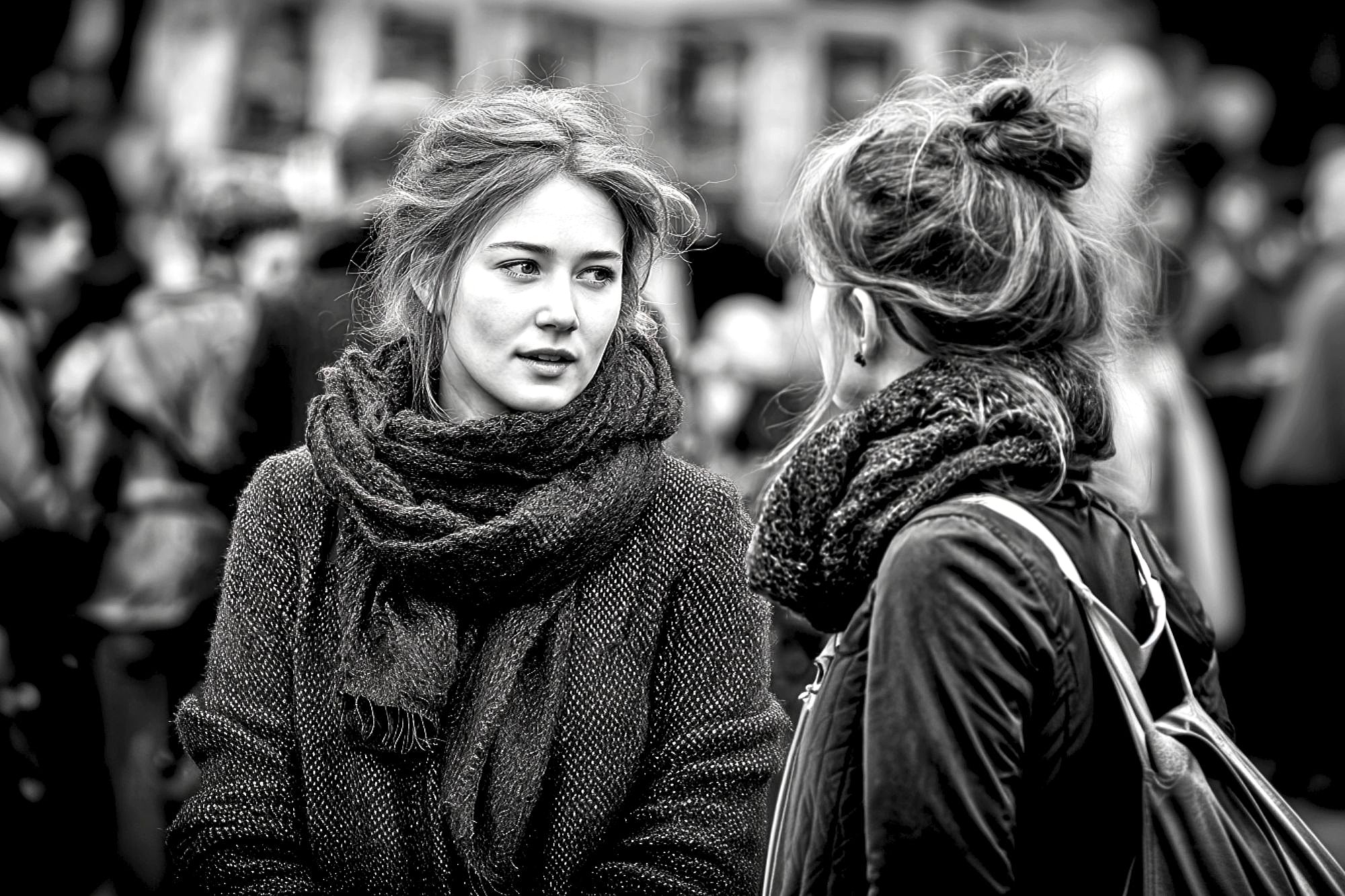 Two young women with scarves talk to each other outdoors in a crowded area. The image is black and white, with a blurred background of people, emphasizing their conversation and expressions.