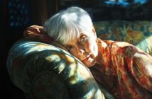 An elderly woman with short white hair rests her head on the arm of a patterned sofa, gazing thoughtfully ahead. She wears a colorful, floral blouse and is illuminated by soft, dappled sunlight.