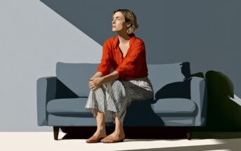 A woman in a red shirt and striped pants sits barefoot on a blue sofa, looking thoughtful. Sunlight creates sharp shadows on the wall and floor, highlighting her face and the minimalist setting.