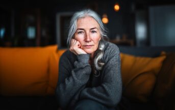 An older woman with long gray hair and a gray sweater sits indoors on a couch with yellow cushions, resting her chin on her hand and looking thoughtfully at the camera. The background is softly blurred.