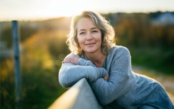 A smiling woman with gray-blonde hair leans on a wooden fence outdoors at sunset, wearing a gray sweater. The background is softly blurred with warm sunlight and greenery.
