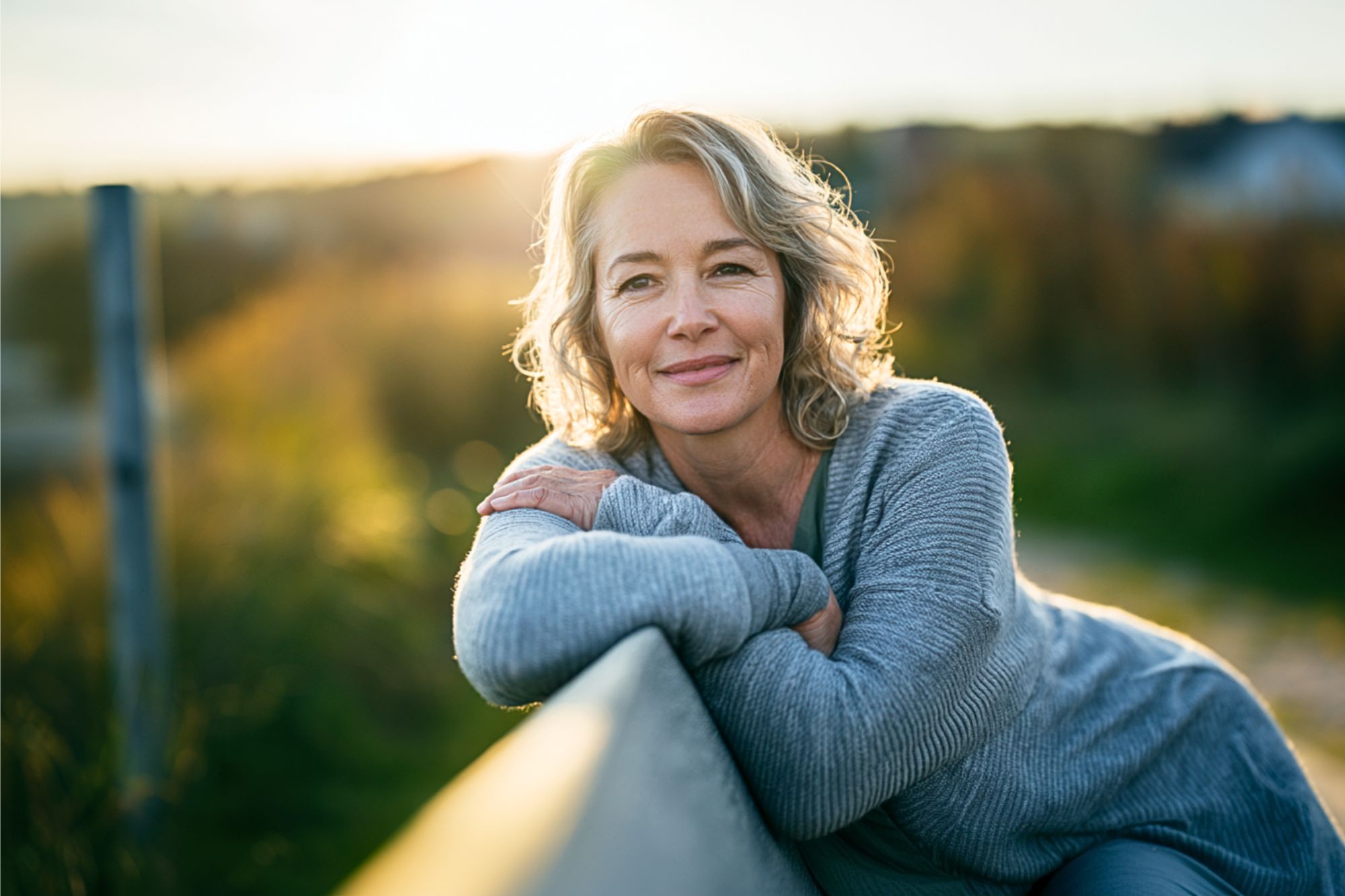 A smiling woman with gray-blonde hair leans on a wooden fence outdoors at sunset, wearing a gray sweater. The background is softly blurred with warm sunlight and greenery.