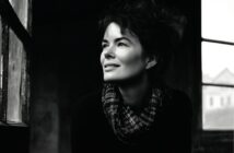 A woman with dark, wavy hair and a checkered scarf smiles as she gazes out a window. The photo is in black and white, creating a soft, moody atmosphere with light illuminating her face.