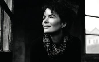 A woman with dark, wavy hair and a checkered scarf smiles as she gazes out a window. The photo is in black and white, creating a soft, moody atmosphere with light illuminating her face.