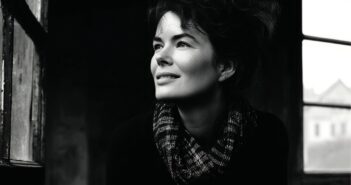 A woman with dark, wavy hair and a checkered scarf smiles as she gazes out a window. The photo is in black and white, creating a soft, moody atmosphere with light illuminating her face.