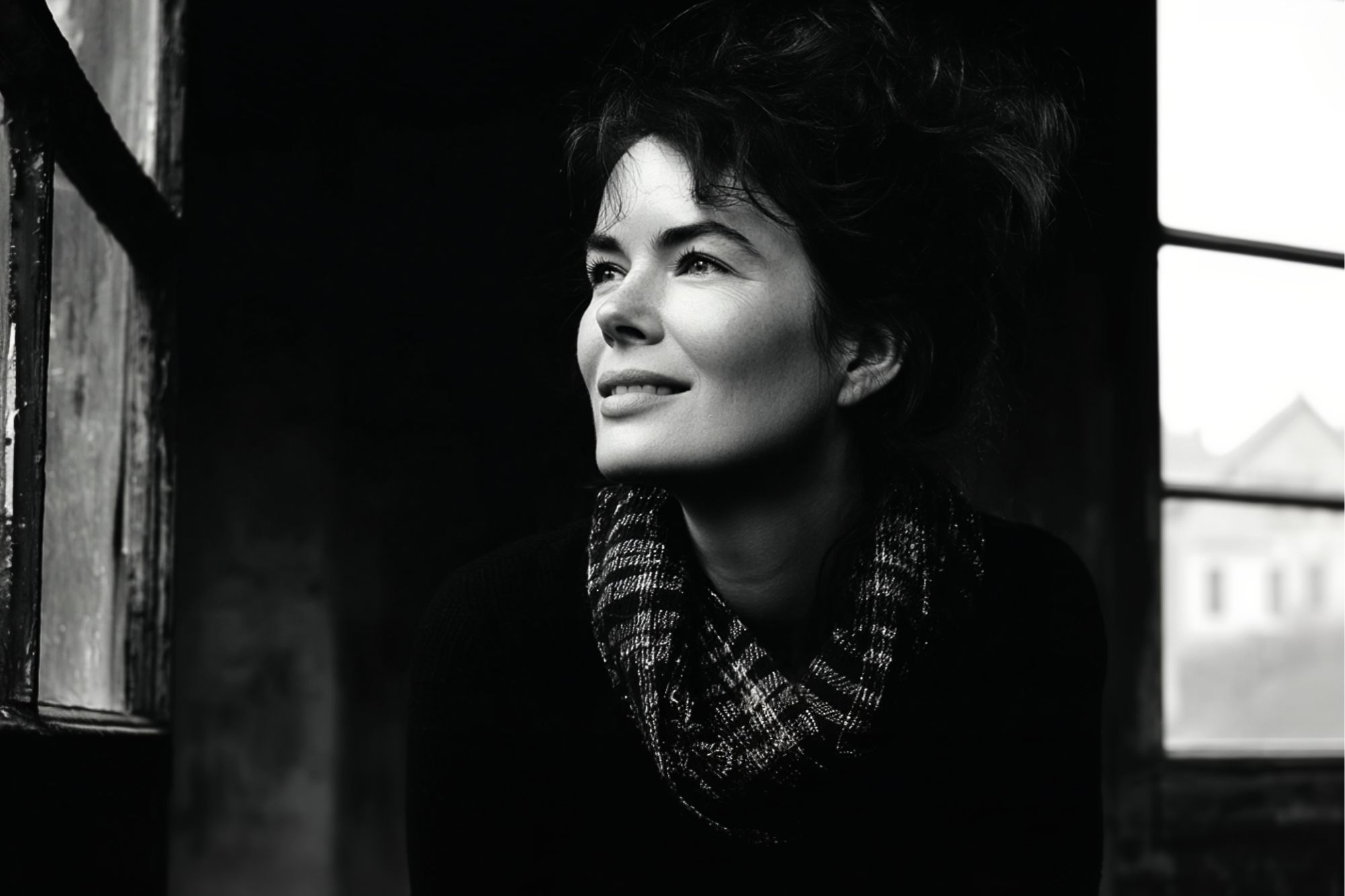 A woman with dark, wavy hair and a checkered scarf smiles as she gazes out a window. The photo is in black and white, creating a soft, moody atmosphere with light illuminating her face.