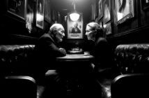 An elderly man and woman sit across from each other at a small table in a dimly lit, narrow cafe lined with framed photographs. They face each other intently, with a cup on the table between them.