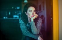 A woman with curly hair and a gray sweater sits indoors, resting her chin on her hand and gazing thoughtfully out of frame. The lighting is soft, with a colorful gradient effect over the image.
