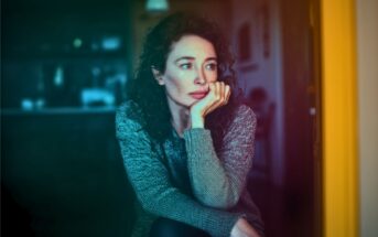 A woman with curly hair and a gray sweater sits indoors, resting her chin on her hand and gazing thoughtfully out of frame. The lighting is soft, with a colorful gradient effect over the image.