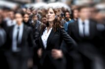 A confident woman in a black suit stands out sharply in focus amid a blurred crowd of people in similar business attire, emphasizing her presence and individuality.