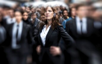 A confident woman in a black suit stands out sharply in focus amid a blurred crowd of people in similar business attire, emphasizing her presence and individuality.