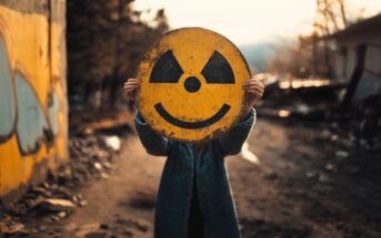 A person stands outdoors on a dirt road, holding a large, round yellow sign with a smiley face and radioactivity symbol in front of their head, obscuring their face. The background is blurred and urban.