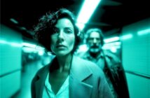 A woman with short curly hair stands in focus under bright fluorescent lights in a corridor, while a man in a jacket stands blurred in the background. The scene has a teal color tone.