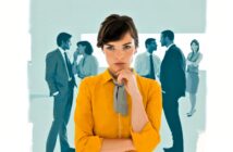 A woman in a yellow shirt stands in the foreground, looking thoughtfully at the camera, while two groups of people in business attire converse in the blurred background.