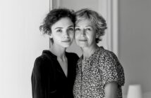 A young woman and an older woman stand close together indoors, looking at the camera with gentle expressions. The image is in black and white, and both women have short, wavy hair.