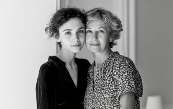 A young woman and an older woman stand close together indoors, looking at the camera with gentle expressions. The image is in black and white, and both women have short, wavy hair.
