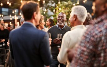 A group of people socializing outdoors at a party or gathering, with string lights overhead. A man in the center smiles while holding a wine glass, engaging in conversation with others.